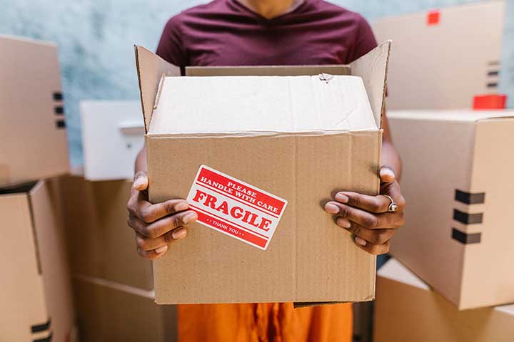 Person carefully holding a moving box labeled 'Fragile', with a stack of other boxes in the background, demonstrating Movers Plus's attention to detail in Frisco.