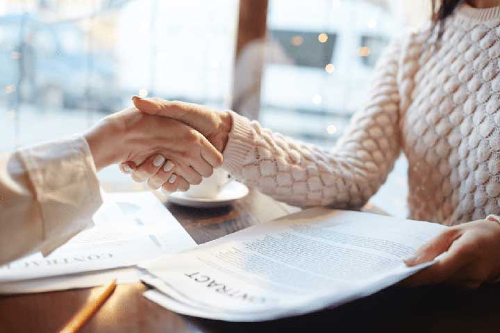 Two individuals shaking hands over a contract in a coffee shop, symbolizing a professional agreement, possibly related to Movers Plus's moving services.
