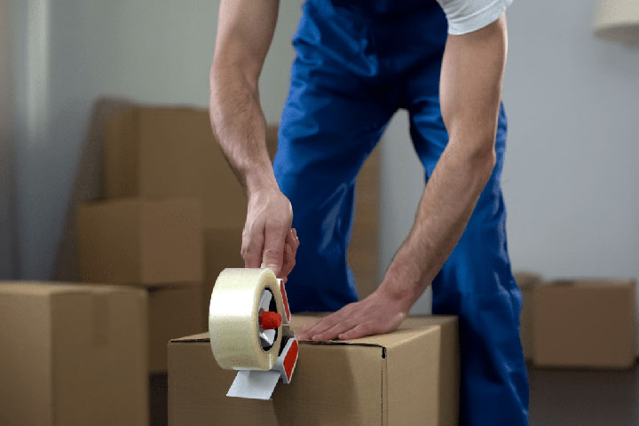 Professional Movers Plus worker sealing a box with tape, surrounded by numerous moving boxes, exemplifying efficient packing services in Frisco.