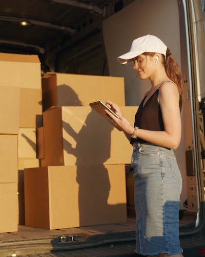 Girl standing in front of a Movers Plus van, surrounded by boxes, attentively checking her iPad to ensure all items are accounted for during the move.