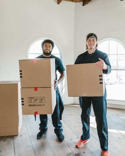 Two smiling movers from Movers Plus standing side by side, holding boxes in an apartment, showcasing reliable and friendly moving services in Frisco.